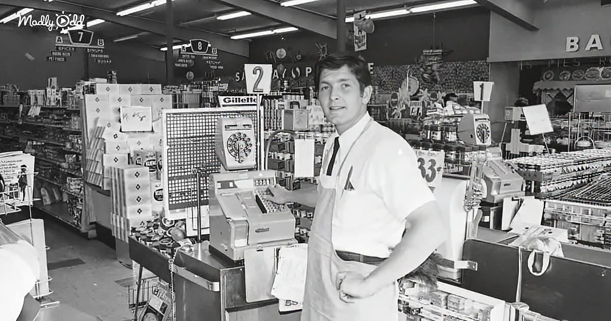 A man in a white shirt and apron stands behind a vintage cash register in a bustling grocery store.