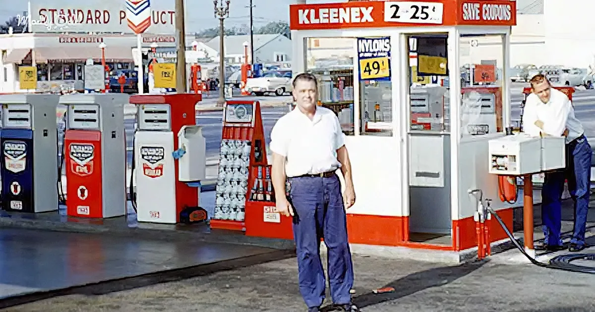 A man in a white shirt stands at a vintage gas station with red and white pumps and a "Kleenex" sign.