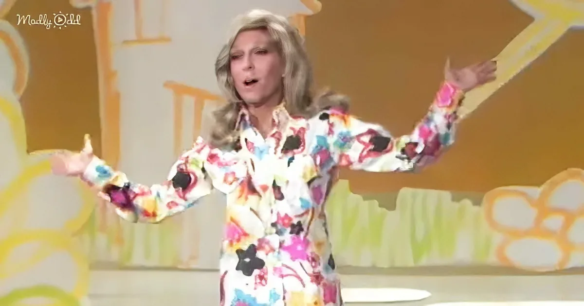 A person in a colorful floral outfit stands with arms outstretched on a stage with a yellow and green background.