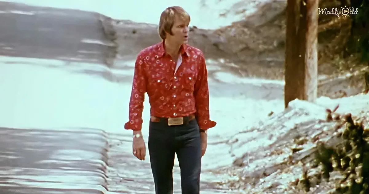 A man in a red floral shirt and jeans walks along a snowy path near a body of water.