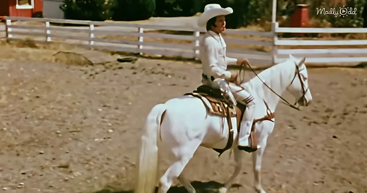 A man in a white cowboy outfit rides a white horse in a fenced arena.