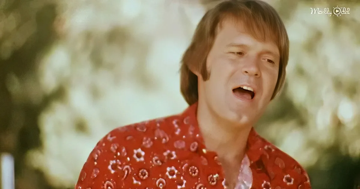 A person in a red patterned shirt is singing with a blurred background of trees and foliage.