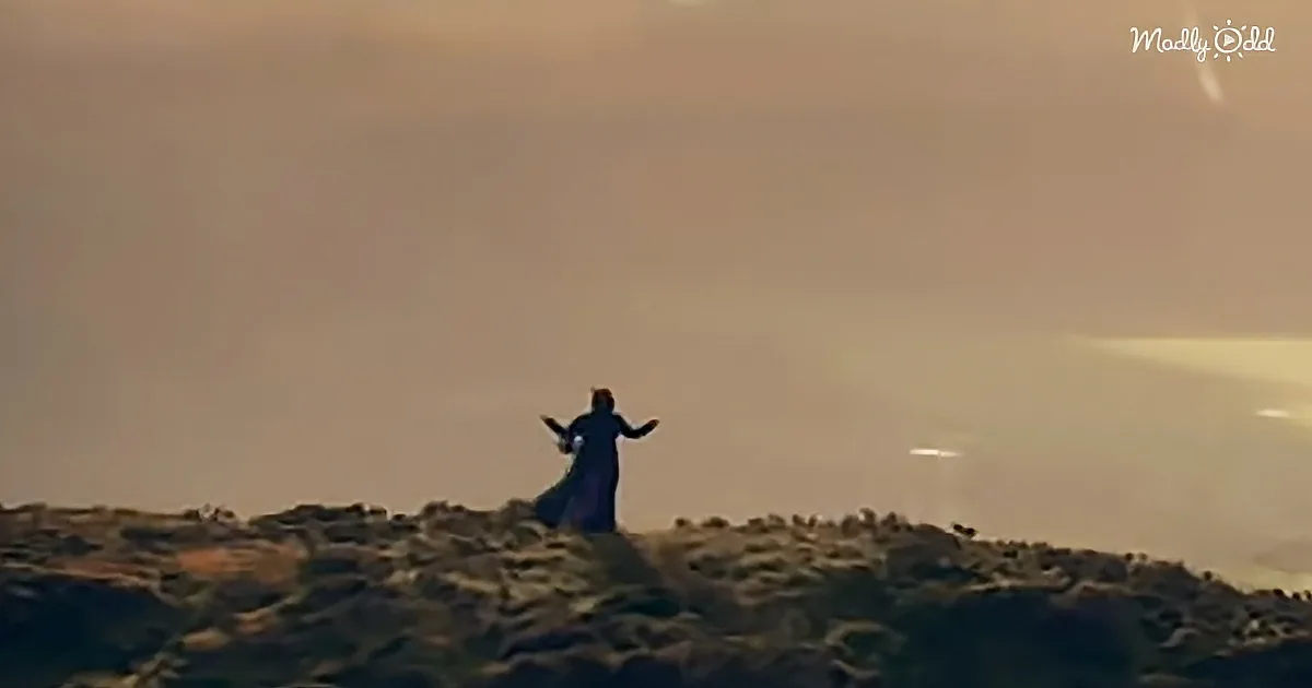 A figure in a dark robe stands on a hilltop, arms outstretched, under a hazy sky.