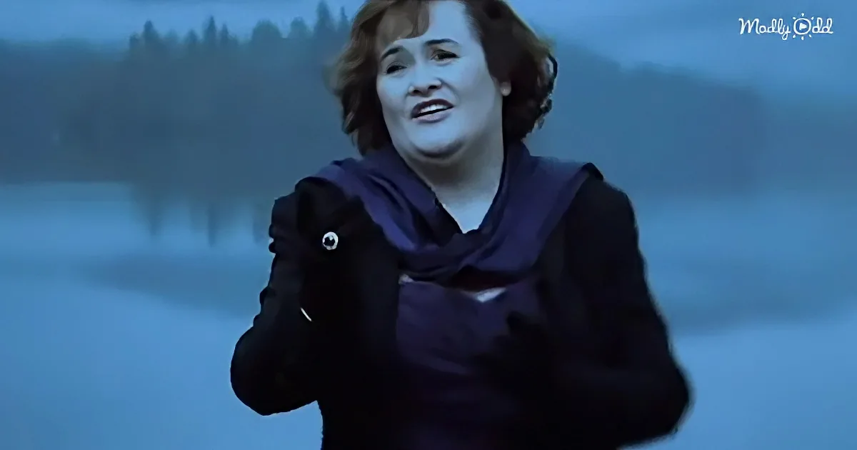 A person in a dark coat and scarf stands outdoors, with a blurred background of trees and water.