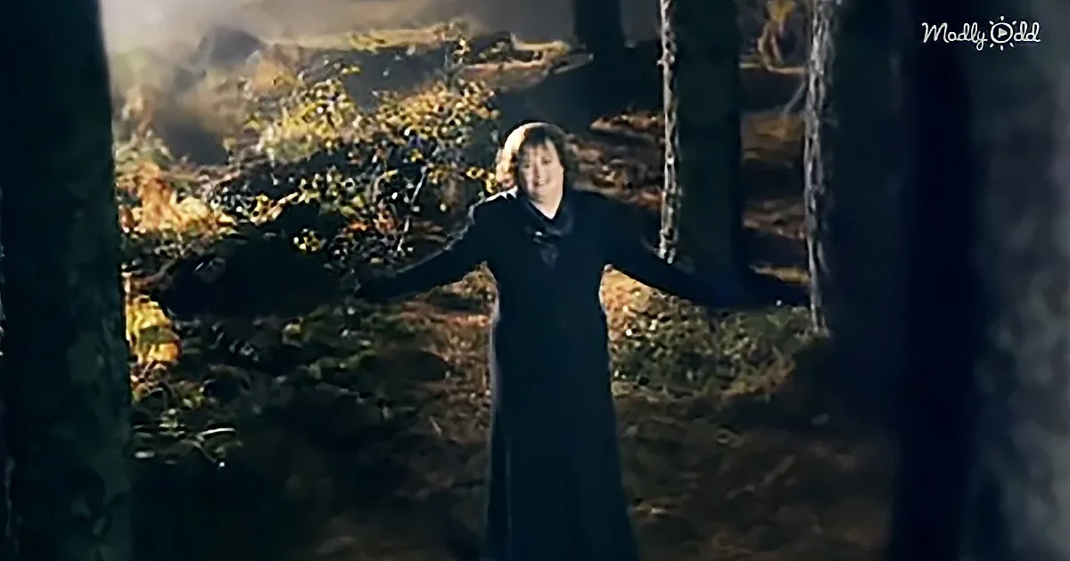 A person in a dark robe stands with arms outstretched in a forested area, surrounded by trees and foliage.