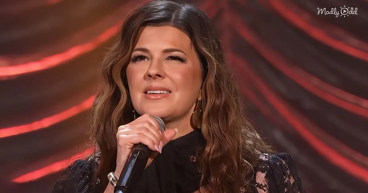 A woman with long, wavy brown hair holds a microphone, wearing a black lace top, against a red and black striped background.