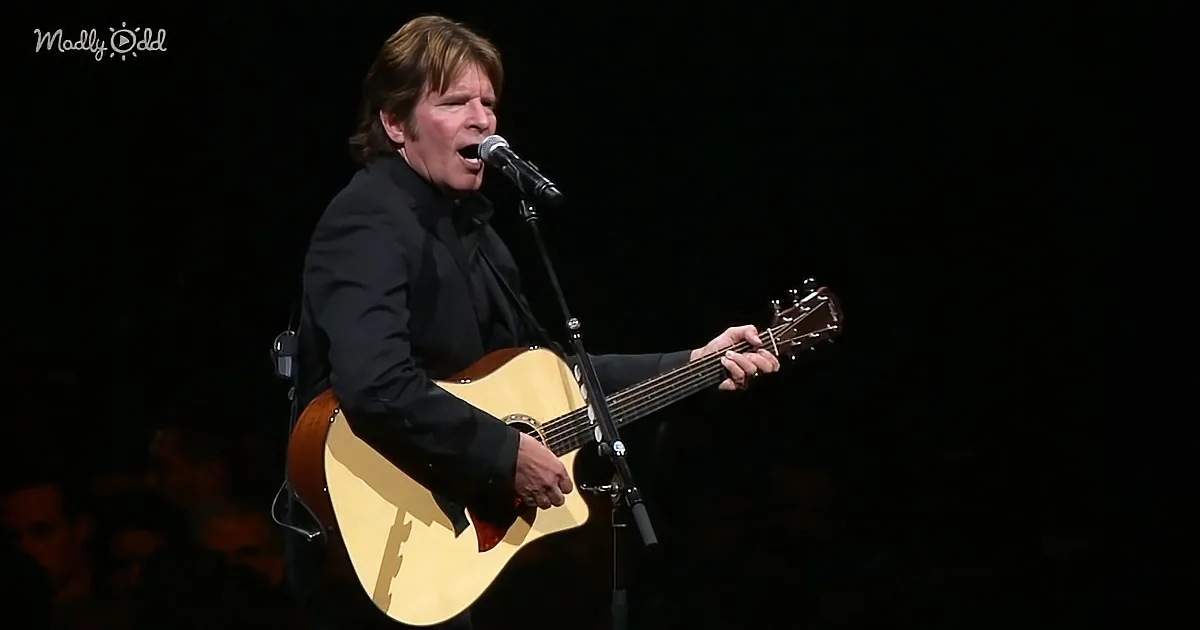 A man in a black suit sings into a microphone while playing an acoustic guitar on stage.