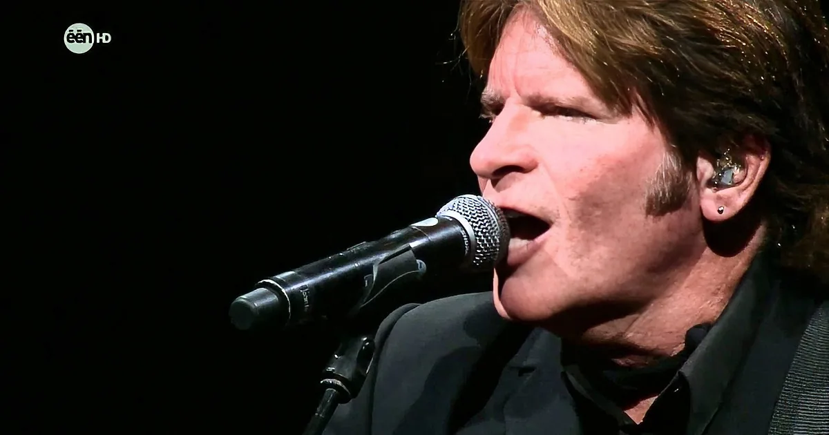 A person singing passionately into a microphone, wearing a black shirt and earpiece, against a dark background.