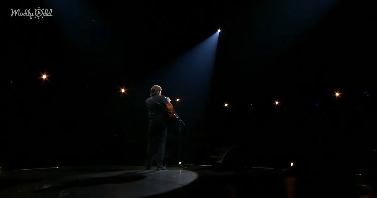 A person stands on stage holding a guitar under a spotlight, surrounded by dark background and scattered stage lights.