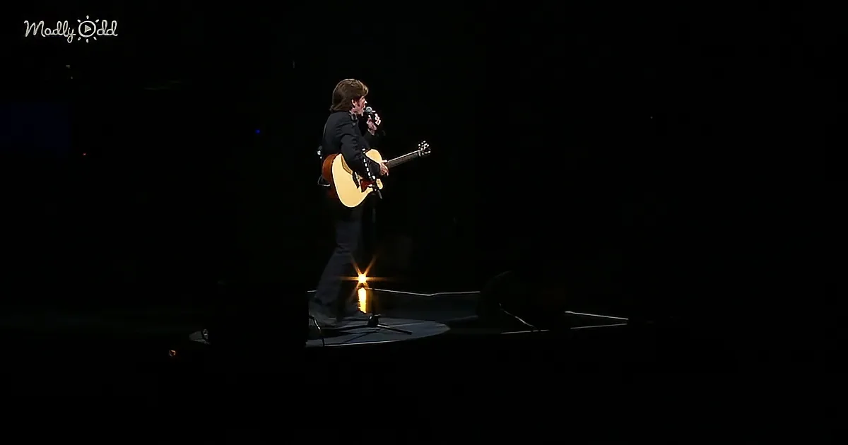 A person stands on stage holding a guitar and microphone, illuminated by a spotlight.