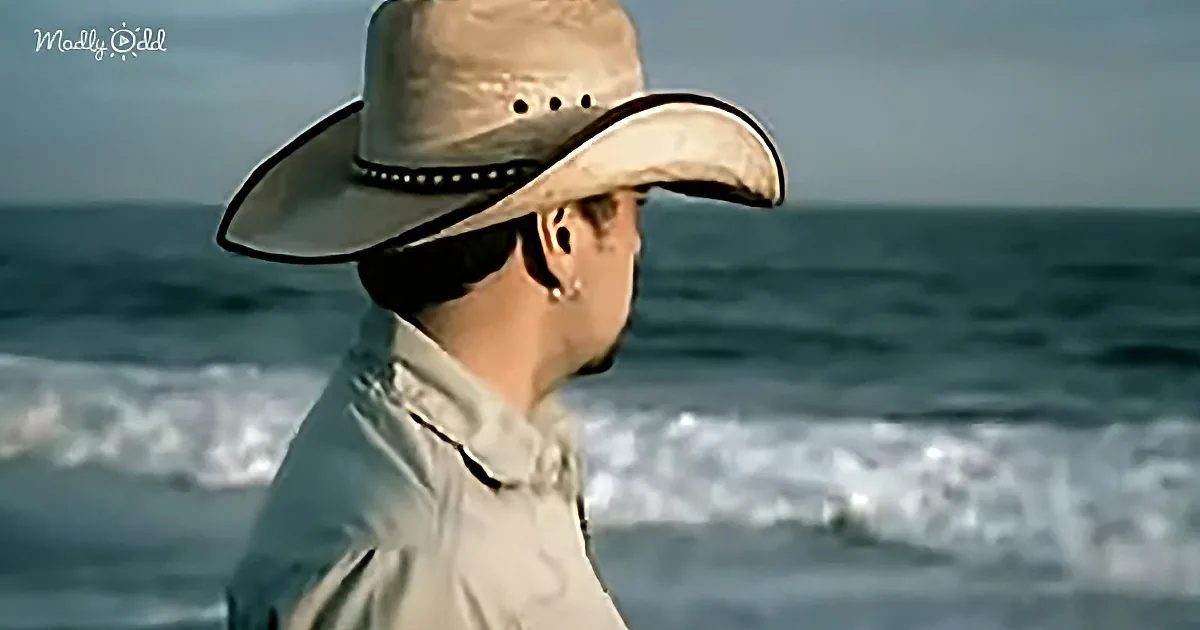 A person wearing a cowboy hat and sunglasses stands by the ocean, gazing out at the waves.