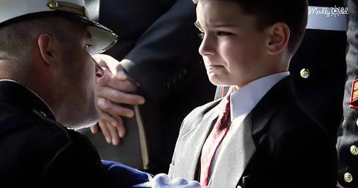A young boy in a military uniform holds a folded flag, while a man in a military uniform leans in, placing a hand on his...