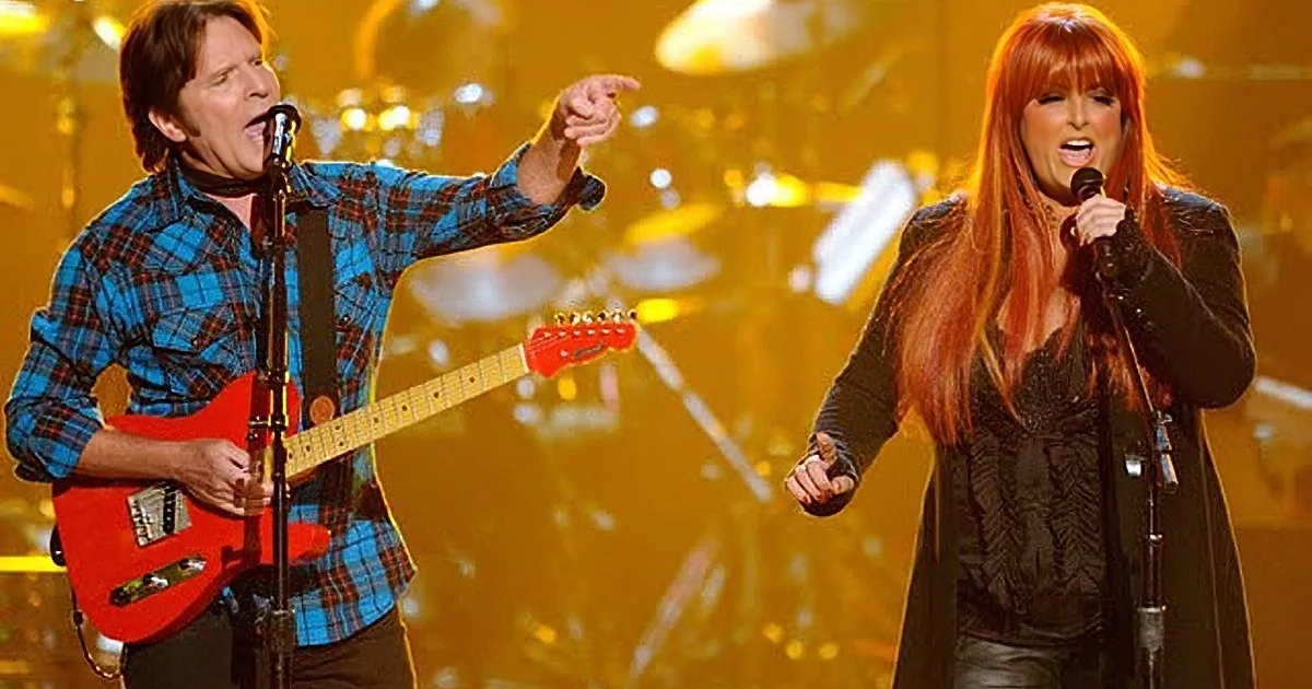 A man plays guitar while a woman sings passionately on stage under warm lighting.