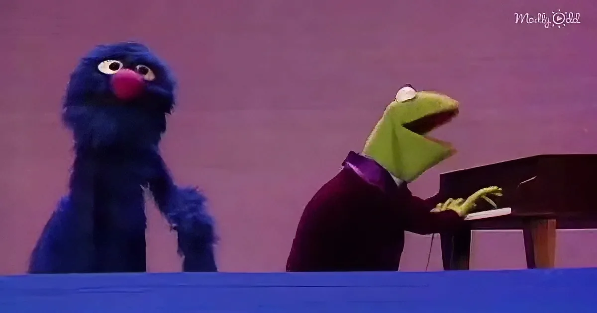 Kermit the Frog plays piano while Grover watches, both puppets on stage.