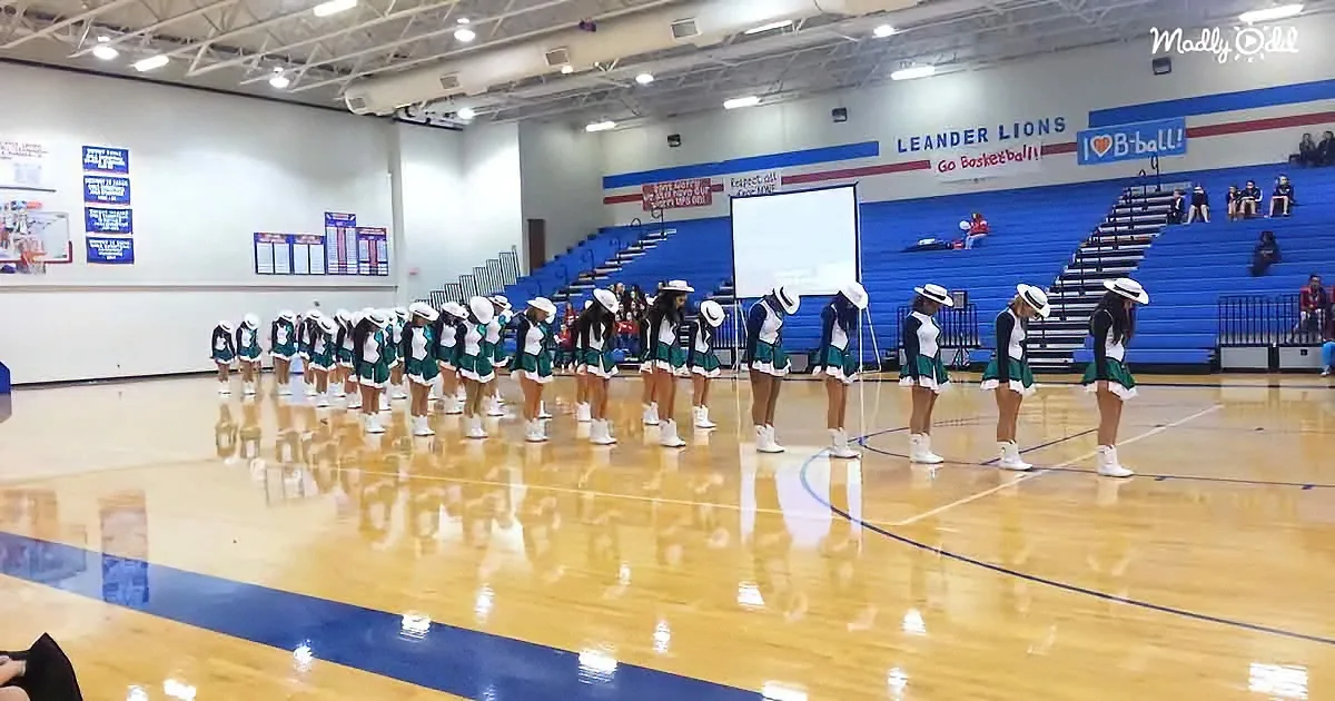 Cheerleaders in green and white uniforms stand in two lines on a polished gym floor.