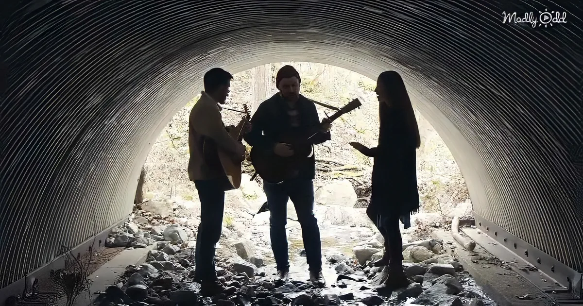 Three musicians play guitars in a tunnel, with a woman standing nearby, silhouetted against the light.