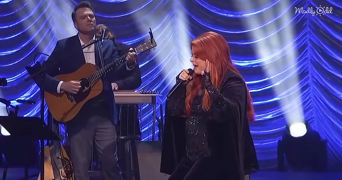 A man plays guitar while a woman sings passionately on stage with blue lighting.