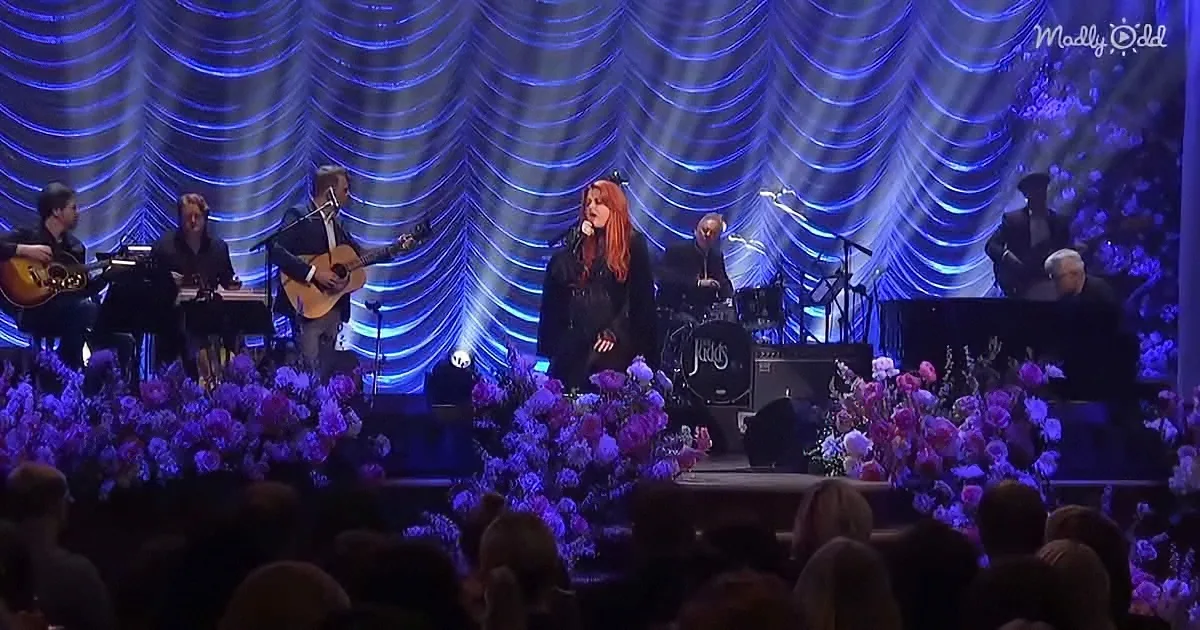 A woman with red hair sings on stage, surrounded by musicians and floral decorations.