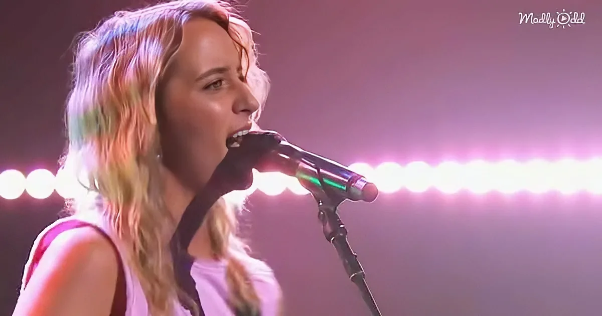 A person with long hair singing into a microphone under bright stage lights.