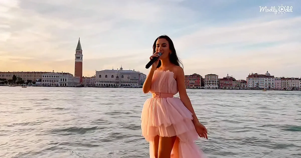 A woman in a pink dress sings into a microphone by the water, with a cityscape and a tall tower in the background.