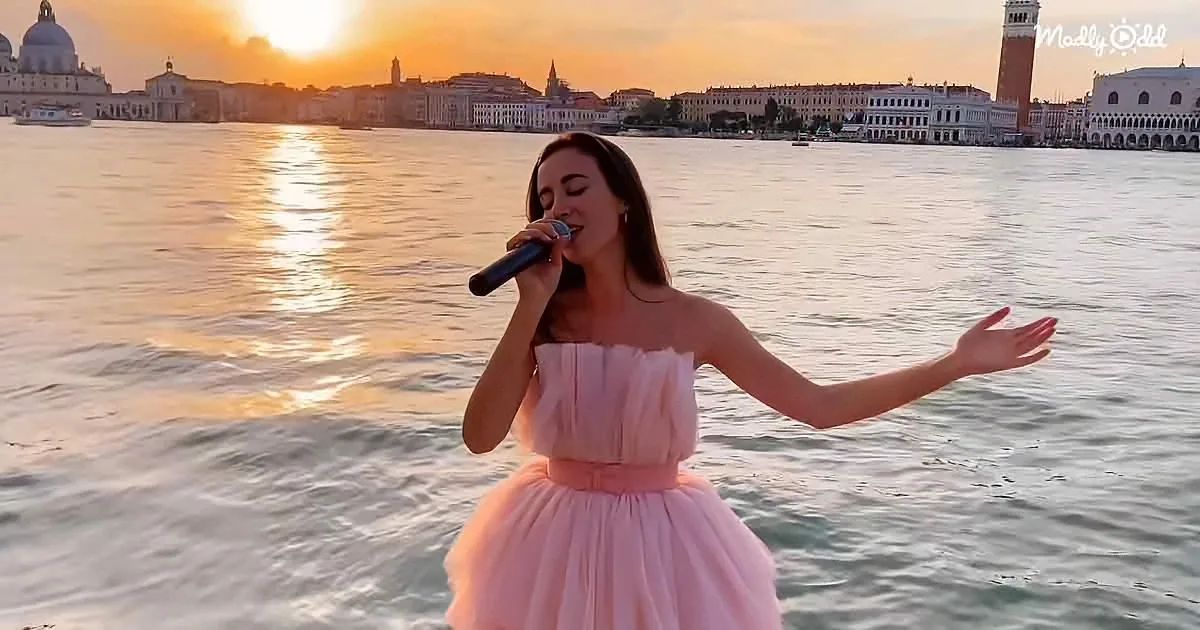 A woman in a pink dress sings into a microphone, standing in front of a serene water backdrop with a cityscape at sunset.