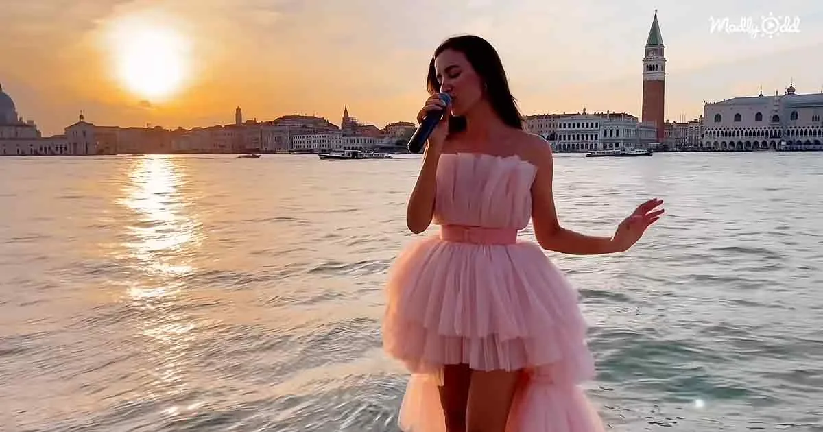 A woman in a pink dress sings into a microphone on a waterfront, with a cityscape and sunset in the background.