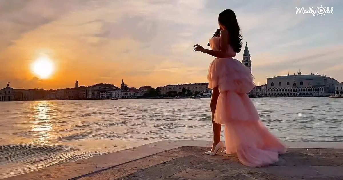 A woman in a pink dress stands on a stone walkway by the water, watching the sunset over a cityscape.