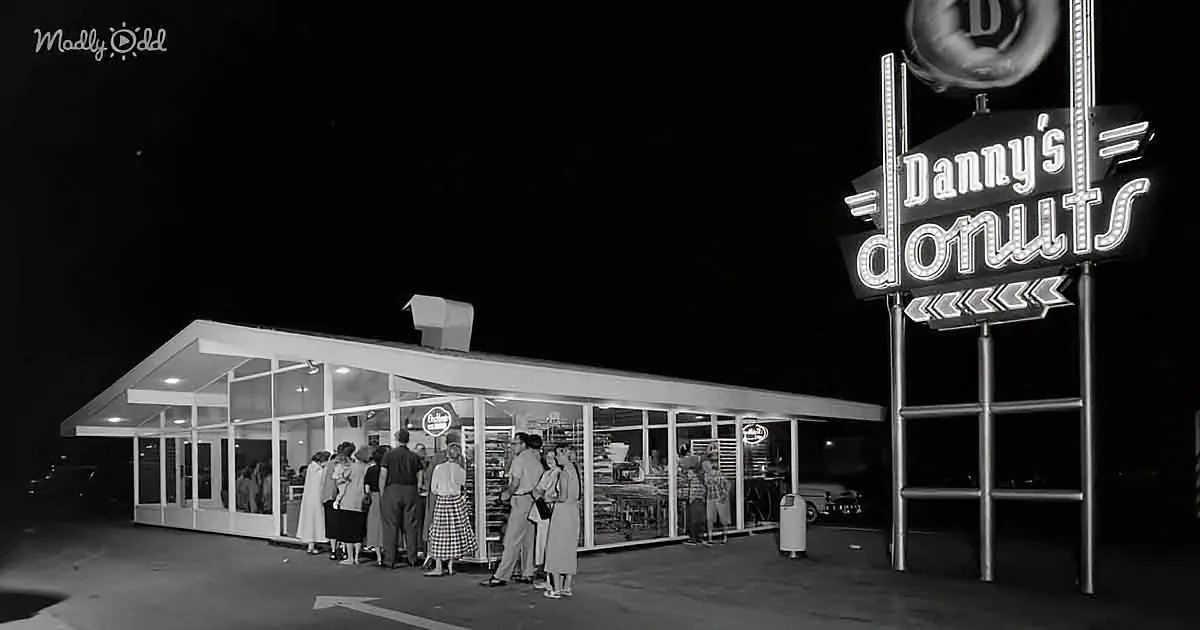 Danny's Donuts neon sign glows brightly at night, attracting a crowd of people outside the illuminated storefront.