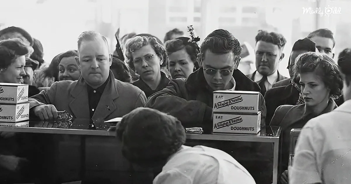 A man in a suit stands behind a counter with Krispy Kreme doughnut boxes, surrounded by a crowd of people.