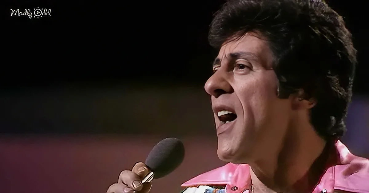 A man in a pink outfit sings into a microphone, his mouth open, with a dark background.