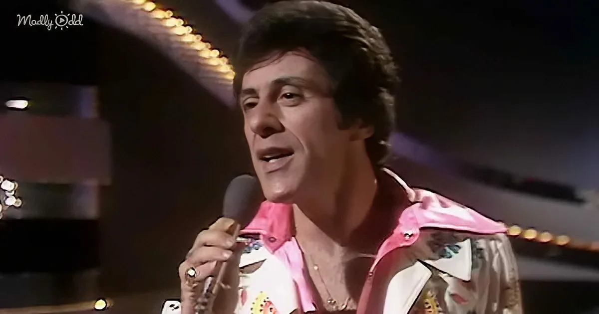 A man in a pink and white shirt sings into a microphone on stage.