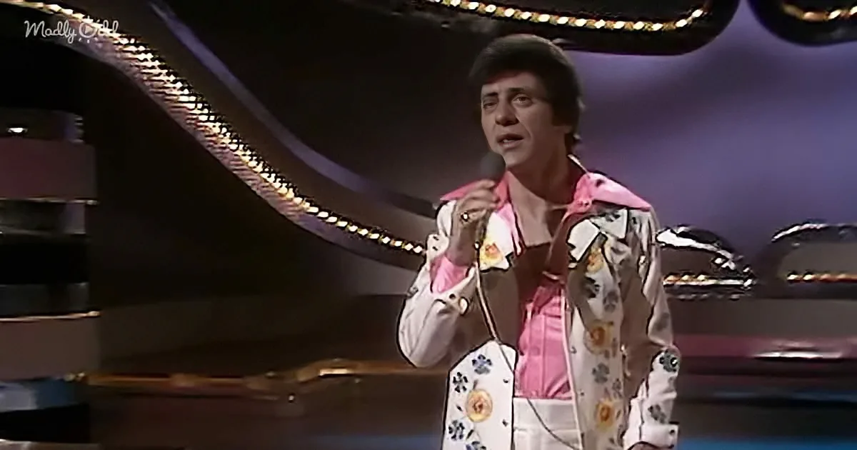 A person in a floral suit sings into a microphone on stage with a piano and lights in the background.
