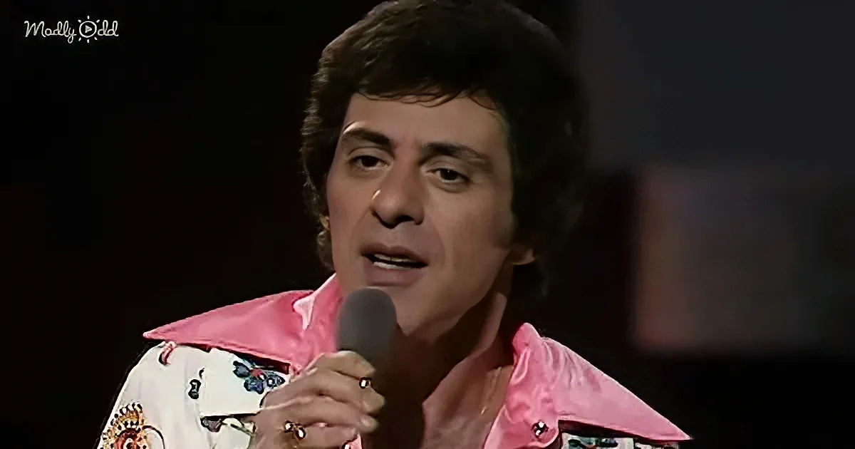 A person in a pink, patterned shirt holds a microphone, possibly singing or speaking on stage.