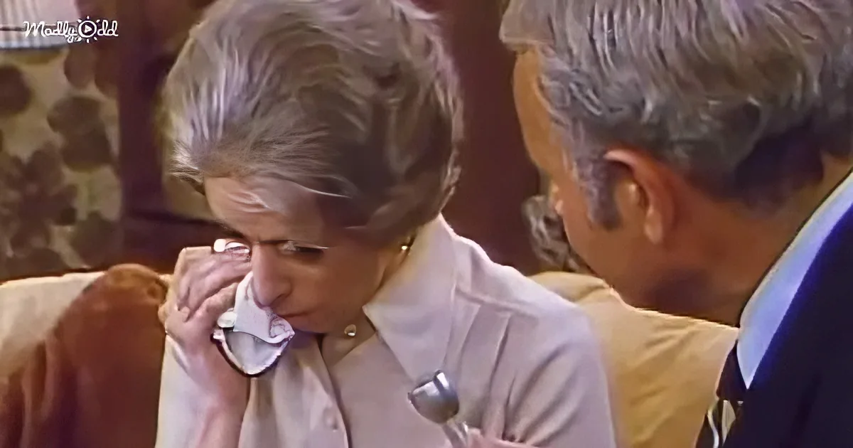 An elderly woman in a white shirt adjusts her glasses while seated, with another person partially visible beside her.