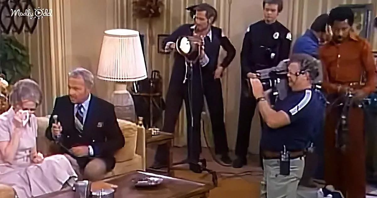 A man in a suit sits on a couch, holding a microphone, while a group of people, including police officers and a...