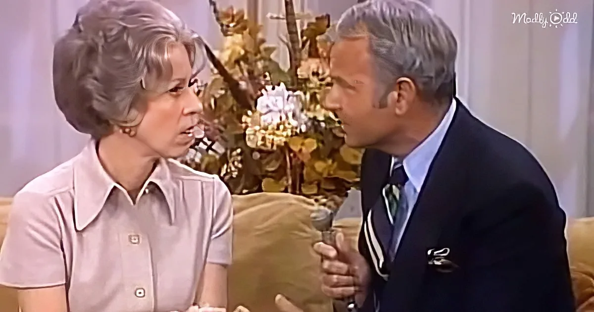 A man in a suit and tie is talking to a woman in a pink shirt, both seated on a couch.