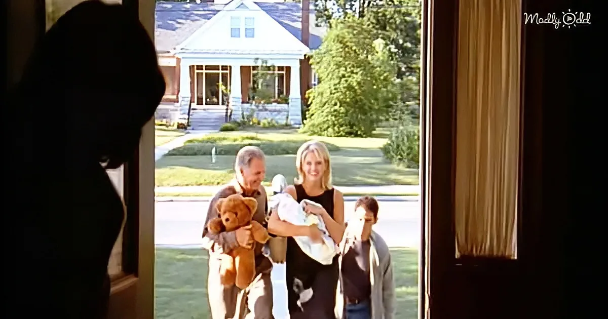 A family with a baby and teddy bear stands outside a house, viewed through a doorway.