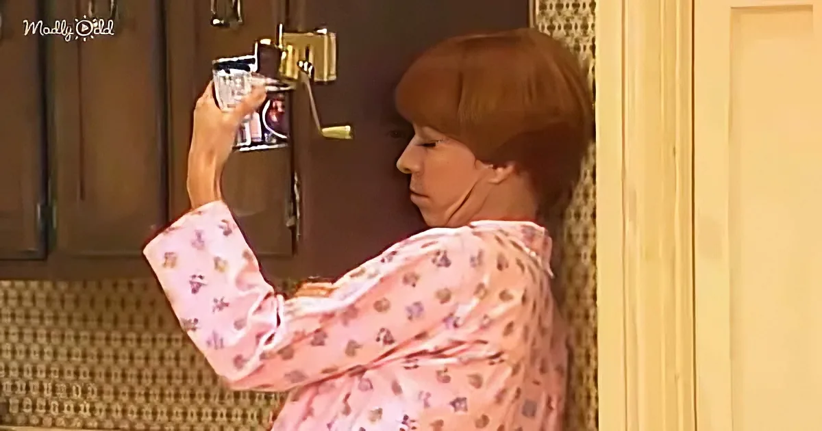 A woman in a pink floral shirt holds a glass and a bottle, looking into a mirror in a kitchen.