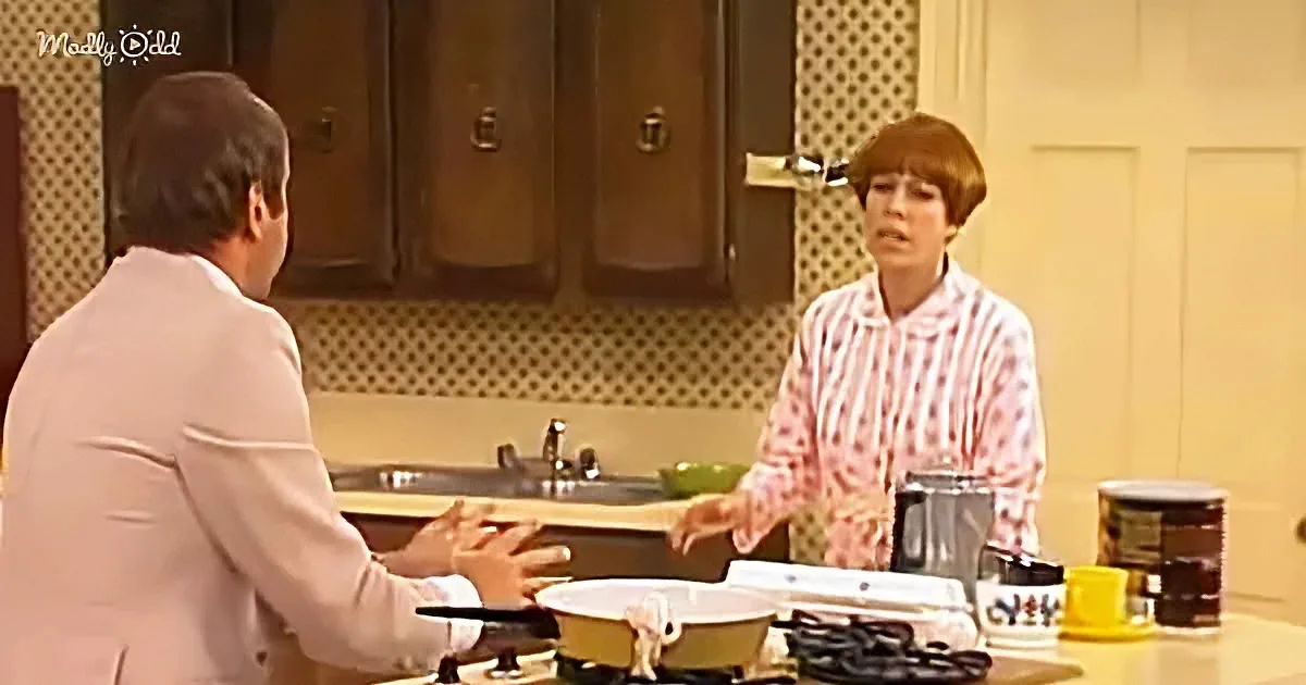A woman in a pink striped shirt stands in a kitchen, gesturing as she speaks to a man in a light suit.