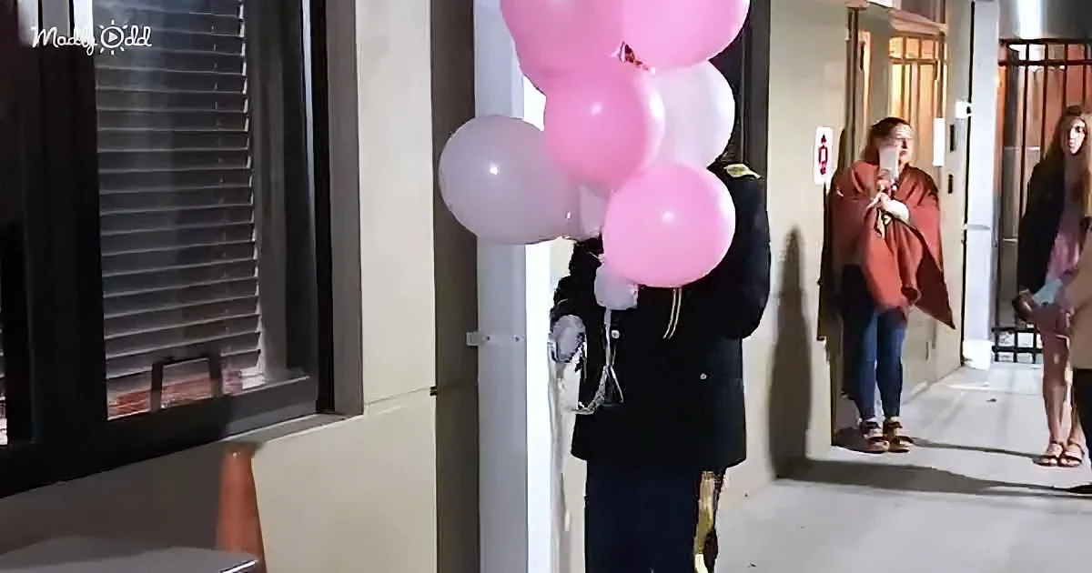 A person in a black coat holds pink and white balloons, standing near a building entrance.