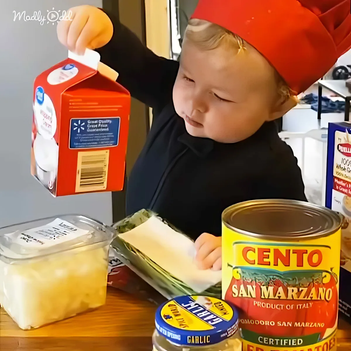 A child in a chef's hat holds a milk carton, surrounded by cooking ingredients on a table.