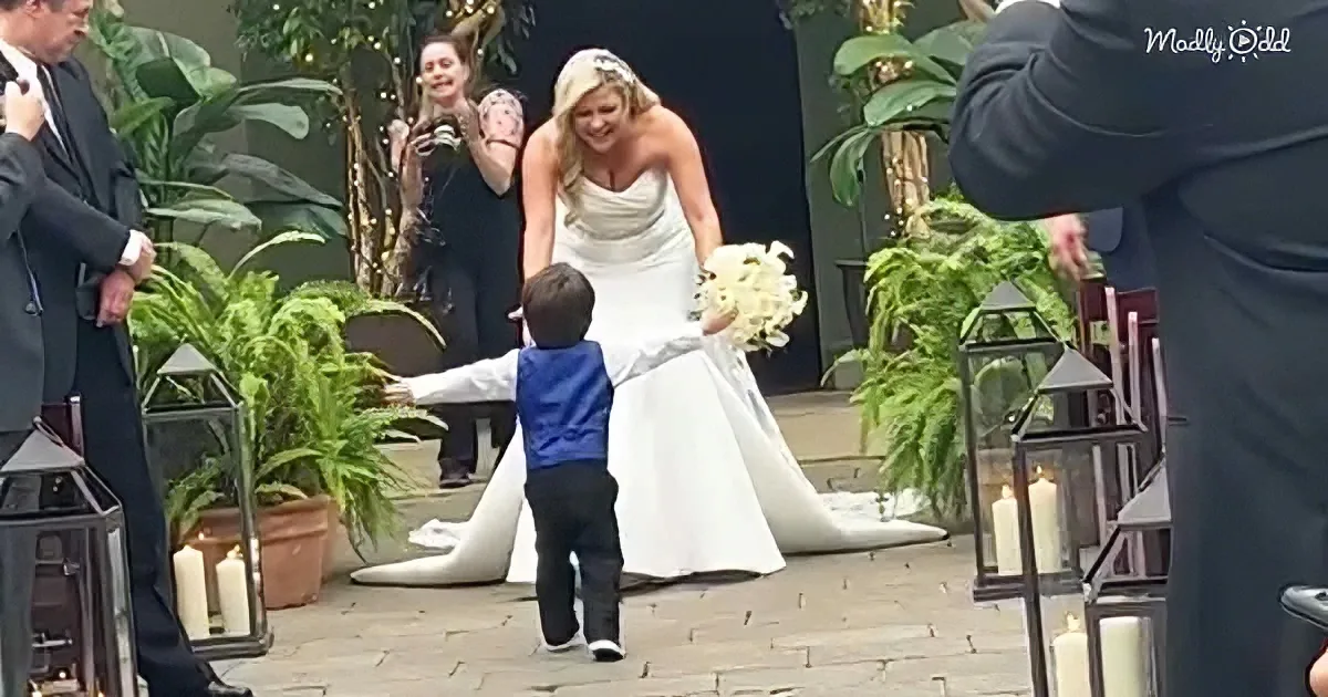 A bride bends down to hug a young boy in a blue vest at a wedding.