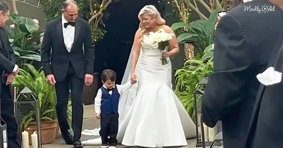 A bride walks down the aisle holding hands with a young boy, surrounded by guests in formal attire.