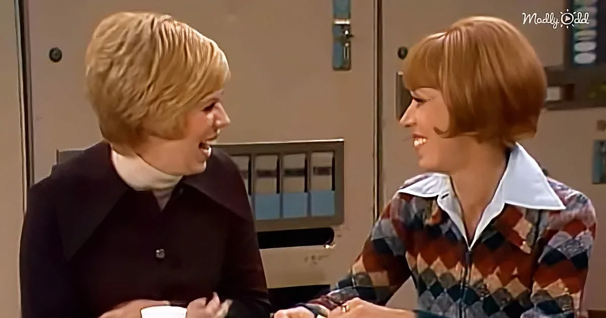 Two women with short hair, one in a dark jacket and the other in a plaid shirt, are smiling and talking in a room.