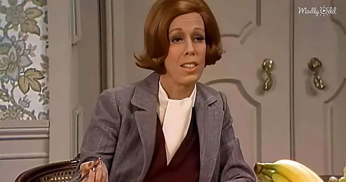 A woman in a gray blazer and maroon top sits in a chair, gesturing with her hands, in a room with floral curtains and a door.