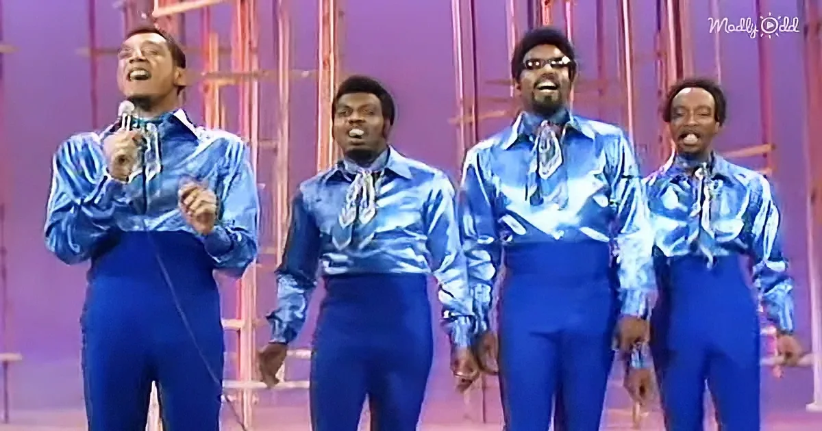 Four men in blue outfits perform on stage, one holding a microphone, against a purple backdrop.