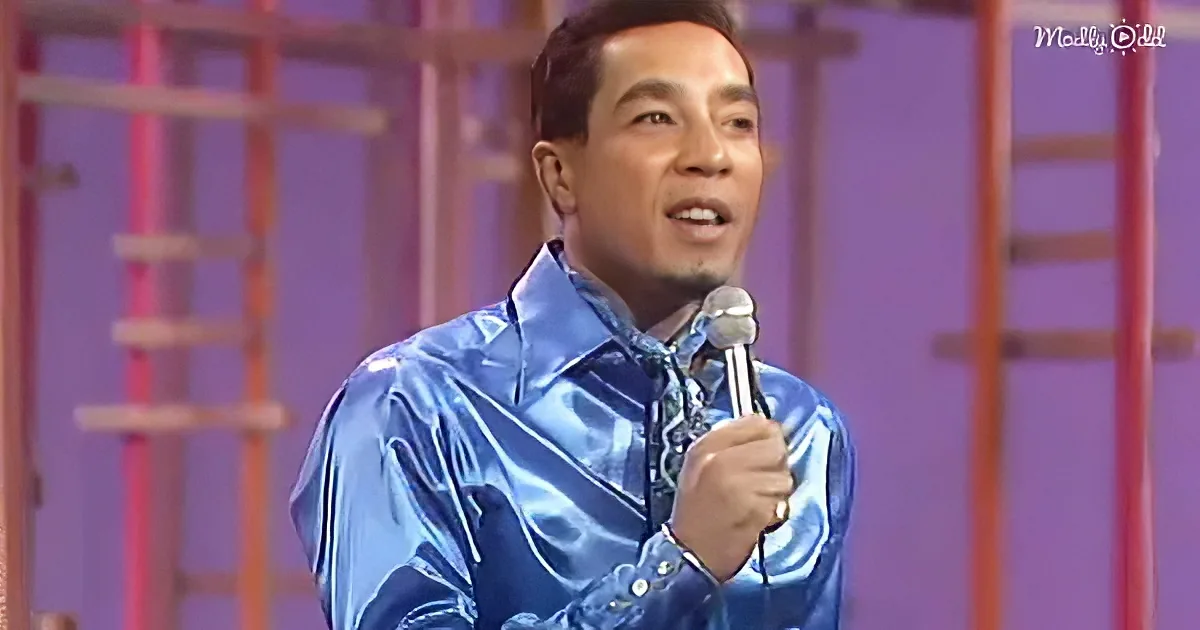 A man in a shiny blue shirt holds a microphone, singing on stage with a purple background.