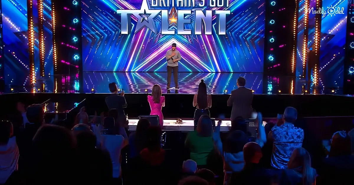 A man stands on a brightly lit stage with "Britain's Got Talent" backdrop, surrounded by judges and an audience.