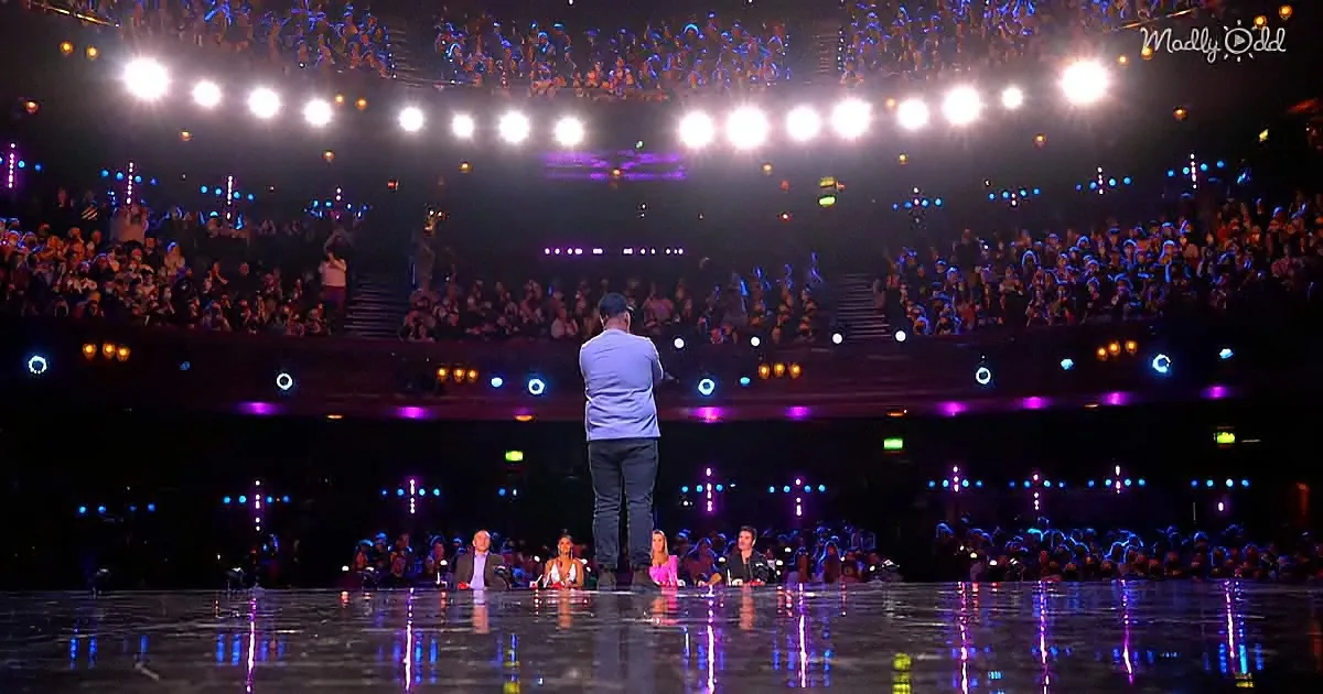 A man stands on stage addressing a large audience in a brightly lit arena.