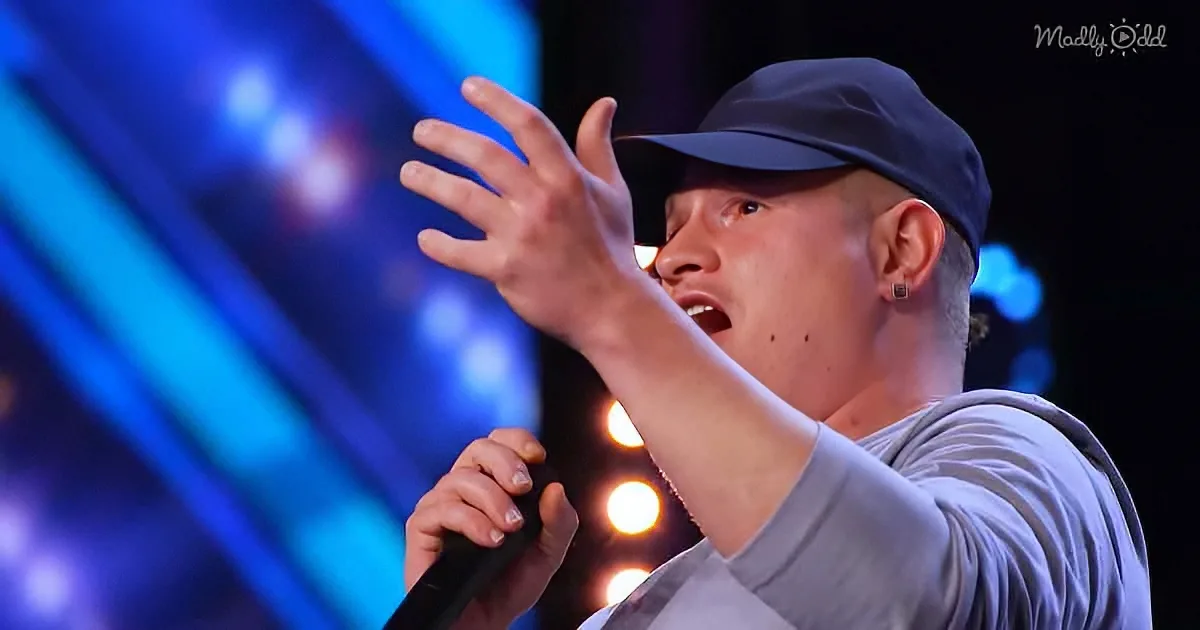 A person in a grey shirt and cap sings into a microphone, raising one hand, with a blue-lit stage in the background.